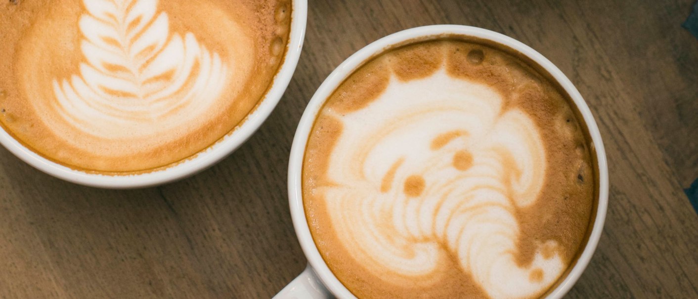 Zwei Cappuccinos mit kunstvoller Latte Art: links ein Blattmuster, rechts ein Elefant, auf einem Holztisch stehend., © Marc Fuhrmann