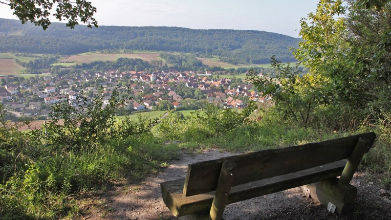 Eine Holzbank bietet einen malerischen Blick auf das Dorf Gündelbach, umgeben von grünen Feldern und bewaldeten Hügeln., © Land der 1000 Hügel - Kraichgau-Stromberg Eine Holzbank bietet einen malerischen Blick auf das Dorf Gündelbach, umgeben von grünen Feldern und bewaldeten Hügeln., © Land der 1000 Hügel - Kraichgau-Stromberg