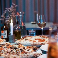 Ein Tisch in einem Restaurant mit mehreren Pizzen, Getr&auml;nken und einer brennenden Kerze. Im Hintergrund sind Gl&auml;ser und eine Flasche zu sehen., &copy; Italo Disco, Stuttgart
