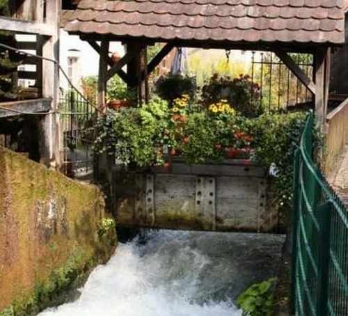 Ein altes Mühlrad mit Blumen geschmückt, umgeben von fließendem Wasser und einem Holzdach., © TOMAS Ein altes Mühlrad mit Blumen geschmückt, umgeben von fließendem Wasser und einem Holzdach., © TOMAS