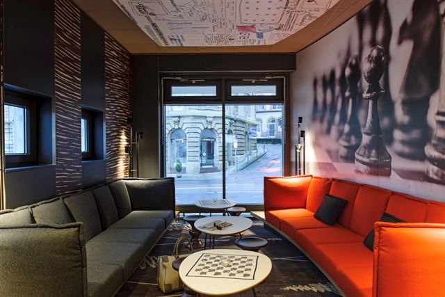 Moderner Raum mit grauen und roten Sofas, Schachbrettmuster auf Tisch, Wand mit Schachfiguren, Fenster mit Blick auf Gebäude., © TOMAS