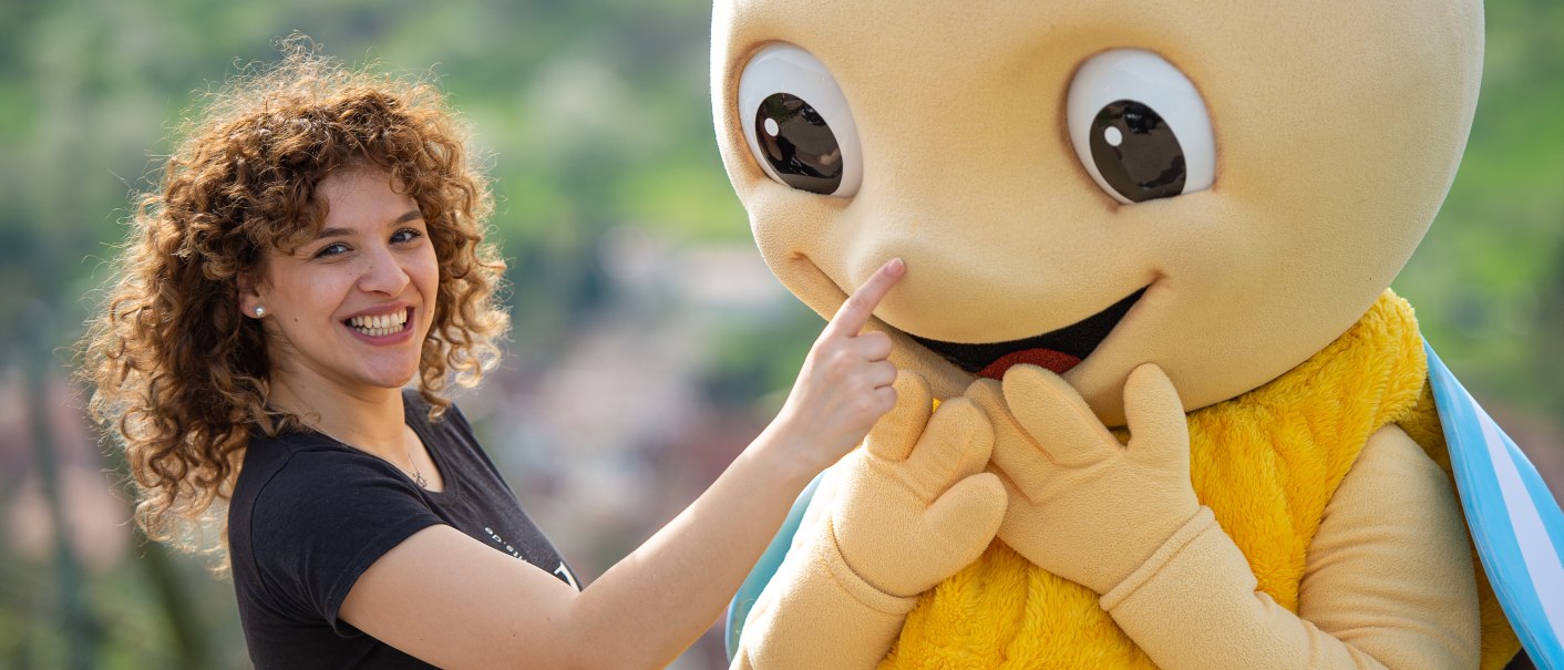 Eine Frau lächelt und berührt die Nase eines großen Bienenkostüms mit gelbem Körper und blauen Flügeln., © Peter Schuster Eine Frau lächelt und berührt die Nase eines großen Bienenkostüms mit gelbem Körper und blauen Flügeln., © Peter Schuster