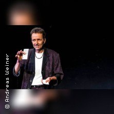 Ein Zauberk&uuml;nstler f&uuml;hrt auf einer B&uuml;hne einen Kartentrick vor. Er tr&auml;gt ein dunkles Sakko und ein wei&szlig;es Hemd., &copy; links im Bild