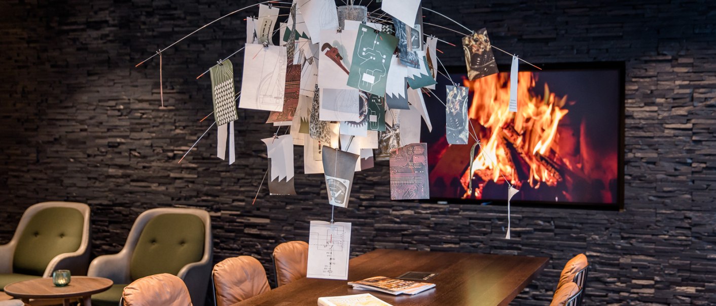 Moderner Raum mit Tisch, darüber hängen Papierblätter an Drähten. Im Hintergrund ein Bildschirm mit Kaminfeuer. Bequeme Stühle und Bücher auf dem Tisch., © TOMAS