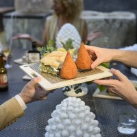 Zwei Personen reichen ein Holzbrett mit Arancini und einem Sandwich an einem Tisch mit mehreren Gästen., © CELEBRE