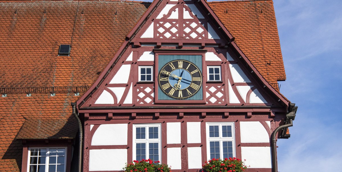 Fachwerk-Rathaus von Bad Urach mit Uhr und Blumenkästen, unter blauem Himmel. Das Gebäude hat ein rotes Dach und eine Turmspitze., © Bad Urach Tourismus