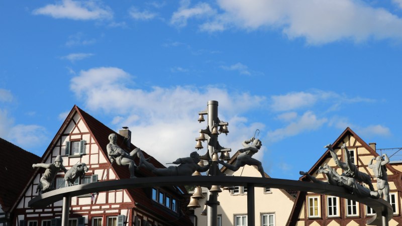 Das Schäferlauf-Denkmal zeigt Figuren und Glocken auf einem Bogen vor Fachwerkhäusern unter blauem Himmel., © Bad Urach Tourismus Das Schäferlauf-Denkmal zeigt Figuren und Glocken auf einem Bogen vor Fachwerkhäusern unter blauem Himmel., © Bad Urach Tourismus