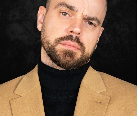 Ein Mann mit Bart und Glatze trägt ein beiges Sakko und einen schwarzen Rollkragenpullover vor einem dunklen Hintergrund., © Scala Kultur Live gGmbH Ein Mann mit Bart und Glatze trägt ein beiges Sakko und einen schwarzen Rollkragenpullover vor einem dunklen Hintergrund., © Scala Kultur Live gGmbH