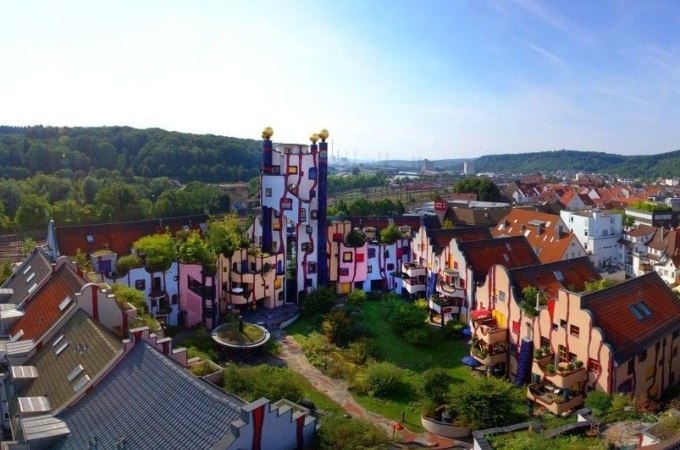 Bunte, unregelmäßige Gebäude im Hundertwasser-Stil mit begrünten Dächern und einem hohen Turm, umgeben von Natur und Stadtlandschaft., © PlochingenInfo