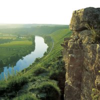 Ein Fluss schlängelt sich durch eine grüne Landschaft, umgeben von Feldern und Hügeln. Im Vordergrund sind Felsen zu sehen., © Land der 1000 Hügel - Kraichgau-Stromberg