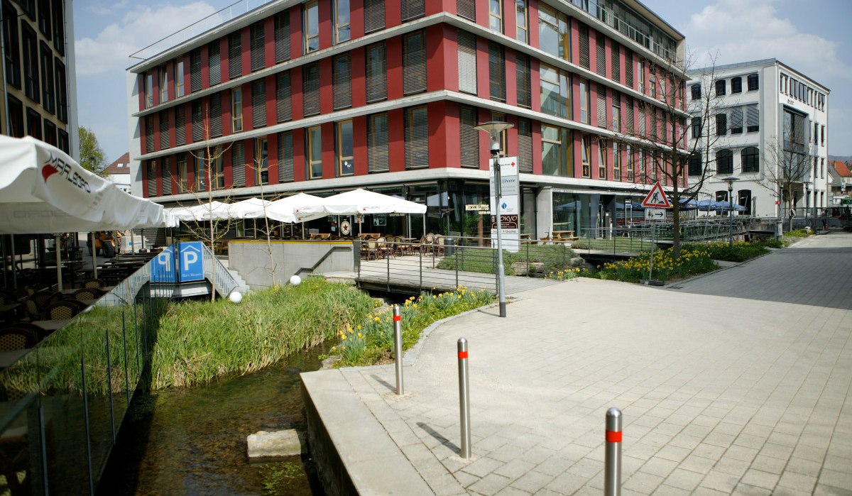 Modernes Gebäude mit roten Fassaden, Glasfronten und Außenterrasse. Ein kleiner Bach fließt daneben, umgeben von Gehweg und Blumen., © Sphäre Verlag