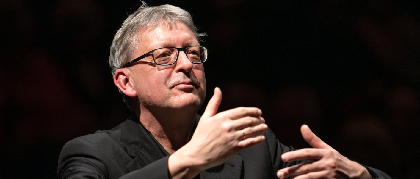 Ein Mann mit Brille dirigiert mit ausdrucksstarken Gesten vor einem dunklen Hintergrund., &copy; Holger Schneider