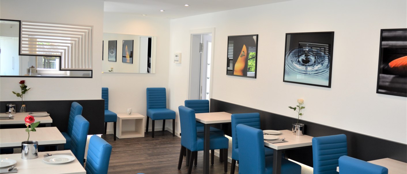 Moderner Frühstücksraum mit blauen Stühlen, weißen Tischen, Blumen und Kunstwerken an den Wänden. Helle Beleuchtung und Holzfußboden., © Hotel BaWü