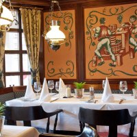 Elegantes Restaurant mit gedecktem Tisch, weißen Servietten, Wandmalerei von Weinbauern und Kronleuchtern. Gemütliche Atmosphäre mit Holzelementen., © Biosphärenhotel Graf Eberhard