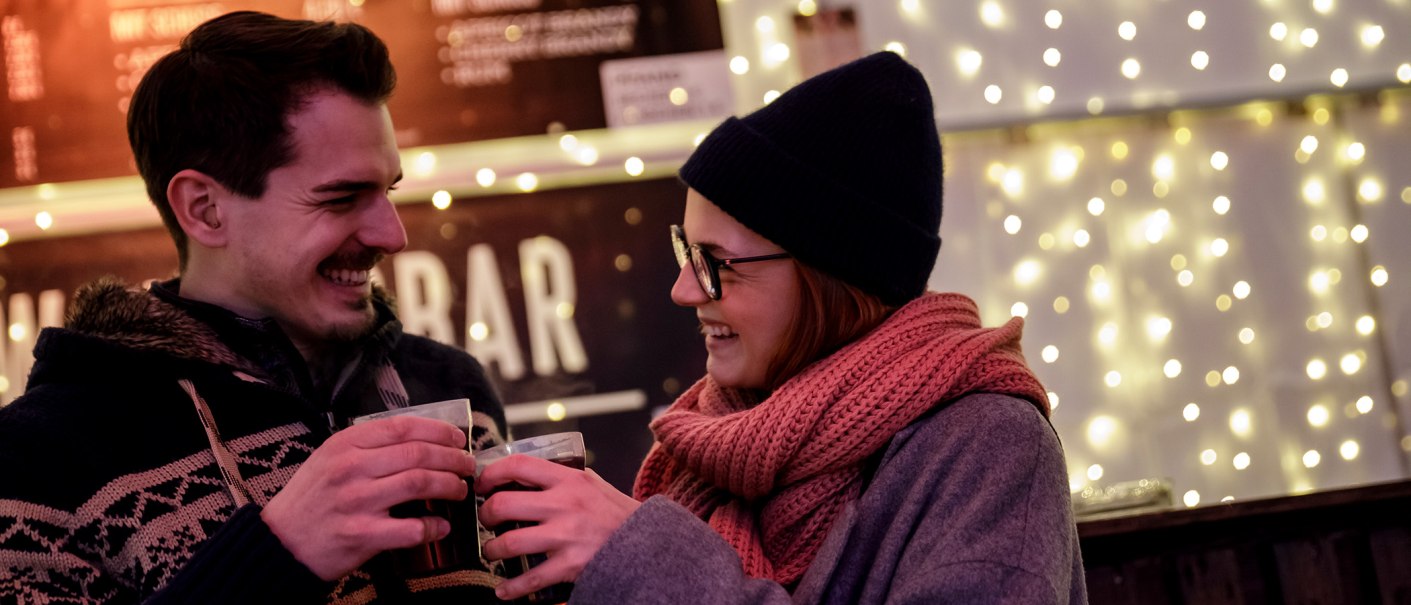 Ein Paar in Winterkleidung stößt mit Glühwein an, umgeben von warmen Lichterketten. Sie lachen und genießen die winterliche Atmosphäre., © Torsten Wenzler Ein Paar in Winterkleidung stößt mit Glühwein an, umgeben von warmen Lichterketten. Sie lachen und genießen die winterliche Atmosphäre., © Torsten Wenzler
