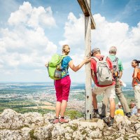Wanderer stehen auf einem Berggipfel neben einem Gipfelkreuz und genießen die Aussicht auf die Landschaft unter einem blauen Himmel mit Wolken., © hochgehberge