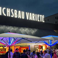 Abendliche Szene vor dem Friedrichsbau Varieté mit beleuchteten Sonnenschirmen und Menschen, die sich unterhalten und entspannen., © Friedrichsbau Varieté Abendliche Szene vor dem Friedrichsbau Varieté mit beleuchteten Sonnenschirmen und Menschen, die sich unterhalten und entspannen., © Friedrichsbau Varieté