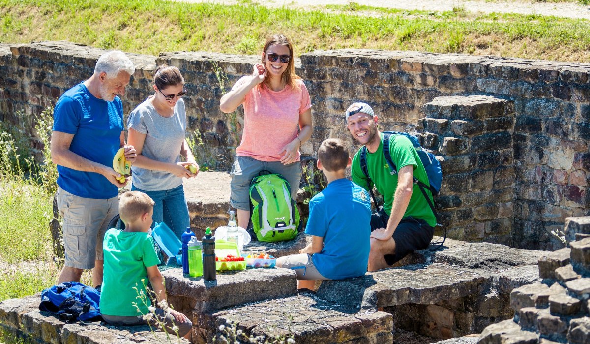 Eine Gruppe von Menschen macht ein Picknick auf alten Steinmauern im Freien. Sie haben Rucksäcke und Essen dabei., © hochgehberge
