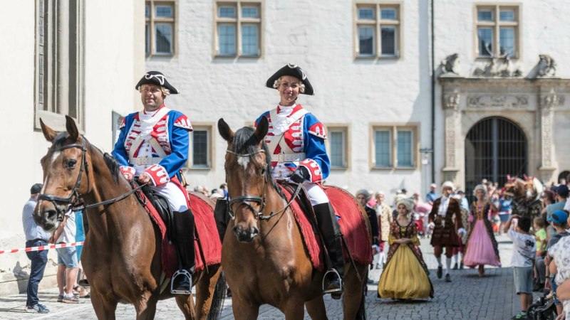 Zwei Reiter in blauen historischen Uniformen auf Pferden bei einem Umzug. Im Hintergrund sind weitere kost&uuml;mierte Personen und Zuschauer zu sehen.