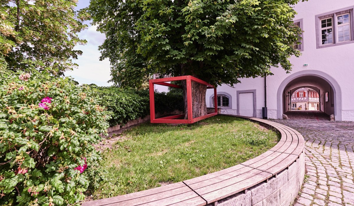 Ein Baum mit rotem Rahmen steht in einem gepflasterten Hof. Im Hintergrund ist ein Torbogen zu sehen. Ein geschwungener Holzboden umgibt den Baum., © Natur.Nah. Schönbuch & Heckengäu Ein Baum mit rotem Rahmen steht in einem gepflasterten Hof. Im Hintergrund ist ein Torbogen zu sehen. Ein geschwungener Holzboden umgibt den Baum., © Natur.Nah. Schönbuch & Heckengäu