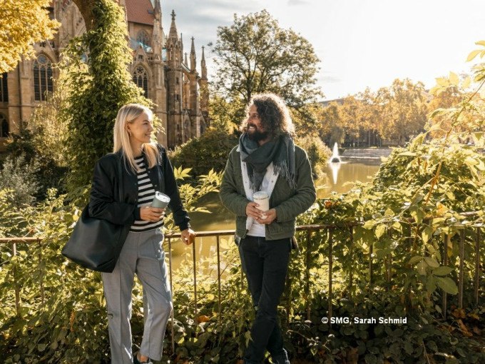Zwei Personen spazieren lächelnd mit Kaffeebechern vor einer Kirche in Stuttgart. Im Hintergrund ein Teich mit Springbrunnen und herbstlicher Vegetation., © Stuttgart Marketing GmbH