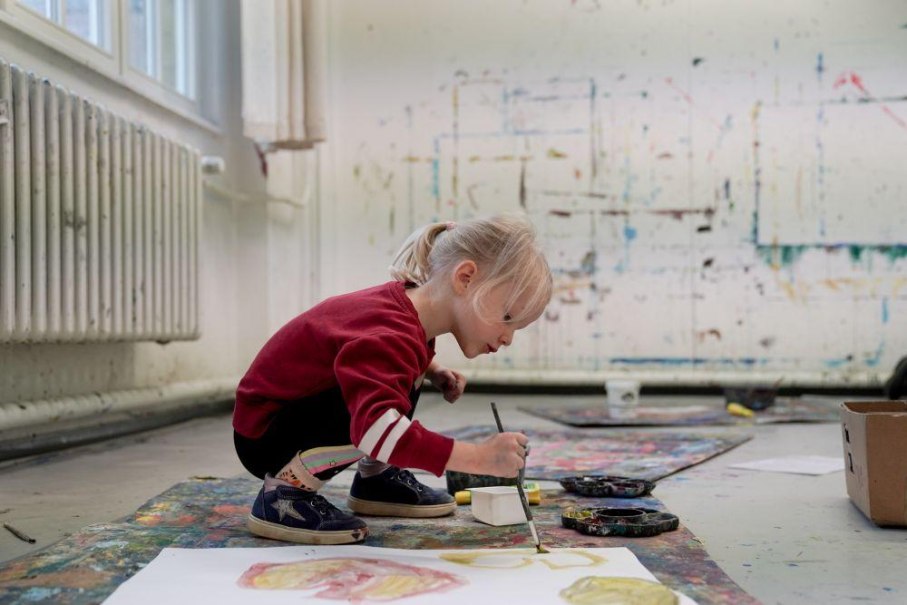 Ein Kind in rotem Pullover malt konzentriert auf dem Boden eines Ateliers mit Wasserfarben. Der Raum ist kreativ gestaltet., © Kunstschule Labyrinth Ein Kind in rotem Pullover malt konzentriert auf dem Boden eines Ateliers mit Wasserfarben. Der Raum ist kreativ gestaltet., © Kunstschule Labyrinth