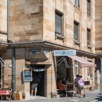 Cafébar Auszeit an einer Straßenecke mit roter Markise. Menschen sitzen draußen, Straßenschilder und Autos sind sichtbar., © SMG, Sarah Schmid