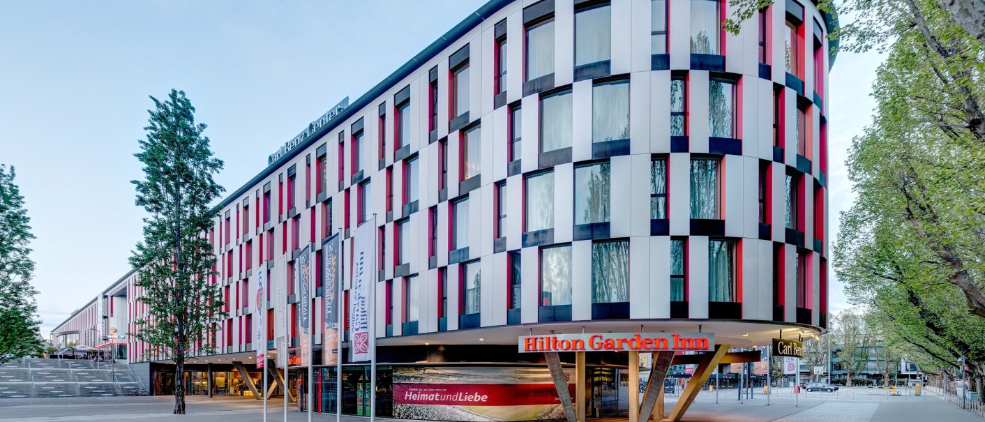 Modernes Hotelgebäude mit bunter Fassade, Hilton Garden Inn Stuttgart Neckarpark. Vor dem Gebäude stehen Fahnenmasten, umgeben von Bäumen., © Hilton Garden Inn Stuttgart Neckarpark Modernes Hotelgebäude mit bunter Fassade, Hilton Garden Inn Stuttgart Neckarpark. Vor dem Gebäude stehen Fahnenmasten, umgeben von Bäumen., © Hilton Garden Inn Stuttgart Neckarpark