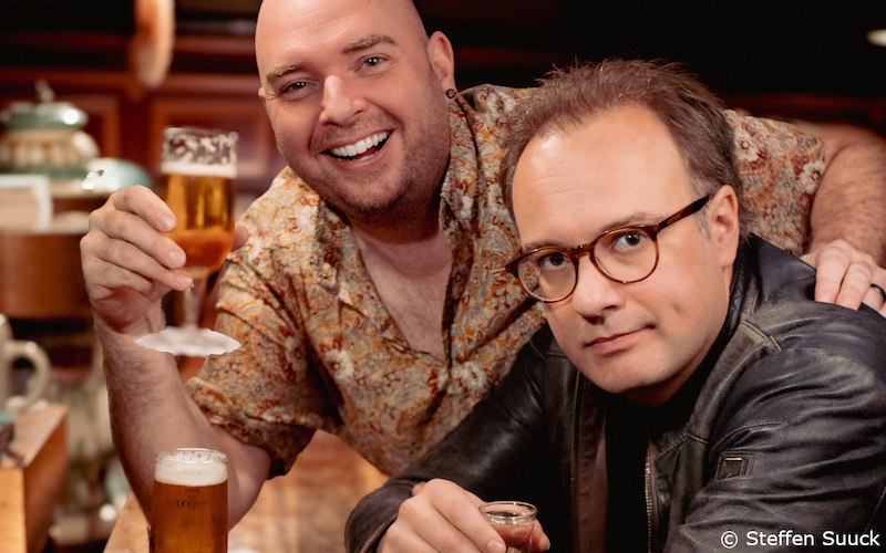 Zwei Männer in einer Bar, einer lächelt und hebt ein Bierglas, der andere schaut ernst und trägt eine Brille und Lederjacke., © Kultur- und Sportamt Stadt Bietigheim-Bissingen