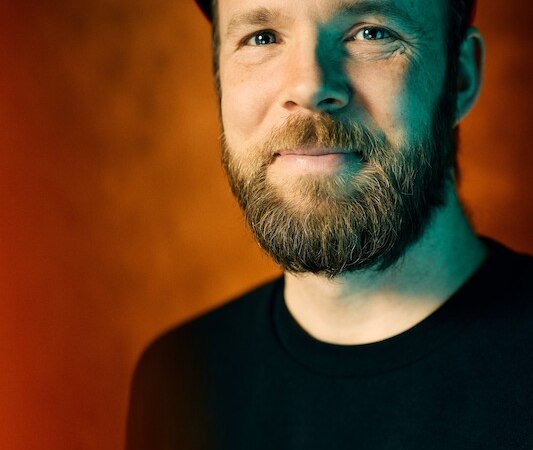 Ein Mann mit Bart und Mütze lächelt vor einem orangefarbenen Hintergrund. Er trägt ein schwarzes Shirt mit einem Smiley-Emblem., © Stadthalle Leonberg Ein Mann mit Bart und Mütze lächelt vor einem orangefarbenen Hintergrund. Er trägt ein schwarzes Shirt mit einem Smiley-Emblem., © Stadthalle Leonberg