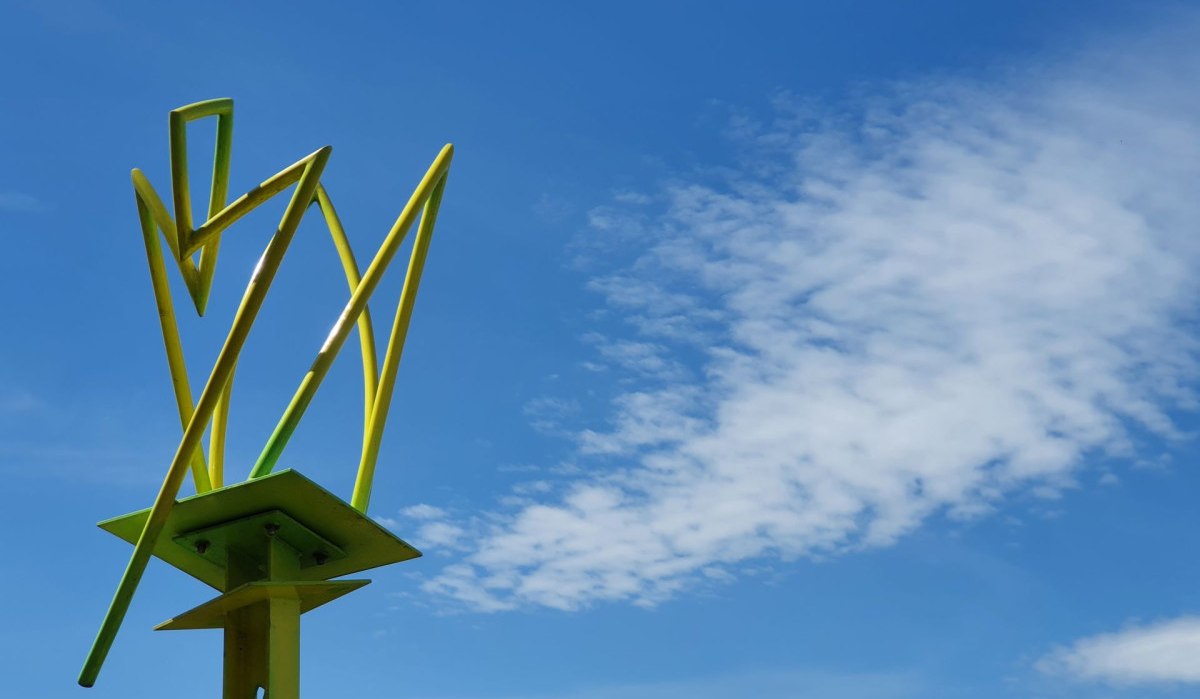 Eine gelbe, abstrakte Skulptur ragt in den blauen Himmel mit einigen Wolken. Im Hintergrund sind Bäume zu sehen.