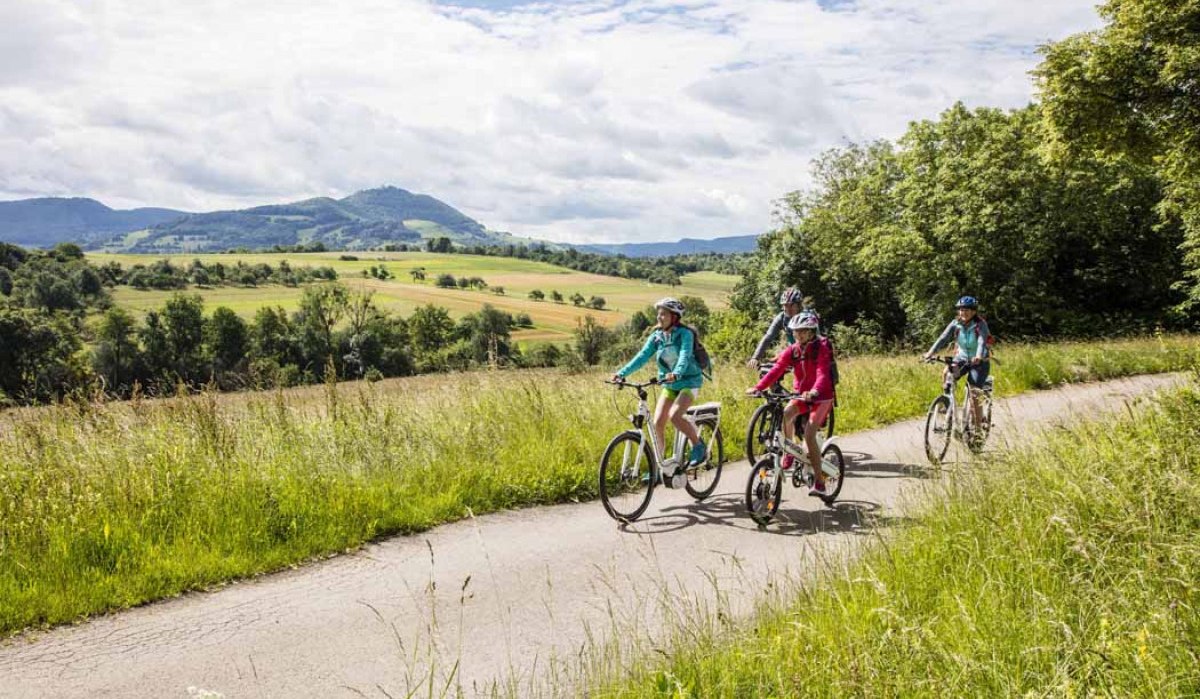 Eine Familie fährt auf einem Radweg durch eine grüne Landschaft mit Hügeln im Hintergrund. Der Himmel ist bewölkt., © Schwäbische Alb Tourismusverband e.V. Eine Familie fährt auf einem Radweg durch eine grüne Landschaft mit Hügeln im Hintergrund. Der Himmel ist bewölkt., © Schwäbische Alb Tourismusverband e.V.