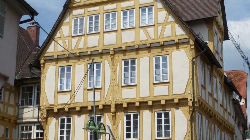 Ein historisches Fachwerkhaus mit gelben Balken, Uhr und Glockenturm. Es steht an einer gepflasterten Straße mit Parkscheinautomat und Blumenkübel.