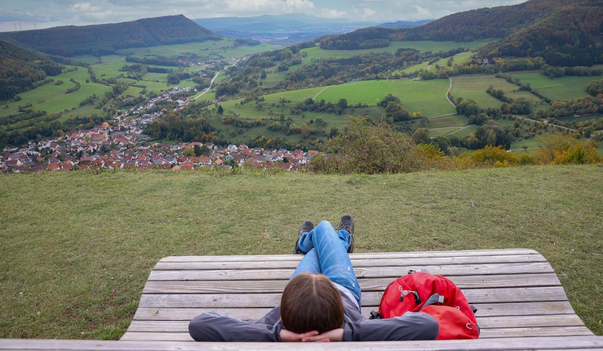 Person entspannt auf Holzbank, blickt auf grünes Tal und Dorf. Hügel und Wälder im Hintergrund, blauer Himmel mit Wolken., © Foto: Mario Klaiber Person entspannt auf Holzbank, blickt auf grünes Tal und Dorf. Hügel und Wälder im Hintergrund, blauer Himmel mit Wolken., © Foto: Mario Klaiber