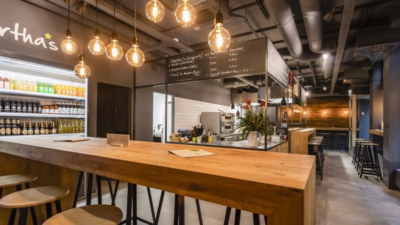 Modernes Café mit Holztischen, Getränkekühlschrank und hängenden Glühbirnen. An der Wand steht "Martha's". Gemütliche Atmosphäre mit Barbereich., © Martha's, Stuttgart