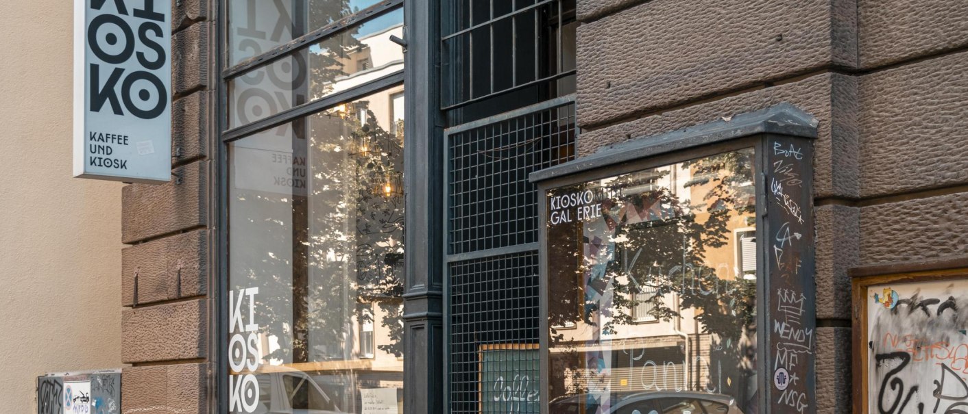 Ein Kiosk mit der Aufschrift 'Kiosko Kaffee und Kiosk'. Die Fassade ist mit Graffiti bedeckt, und das Schaufenster spiegelt die Umgebung wider., © SMG, Sarah Schmid Ein Kiosk mit der Aufschrift 'Kiosko Kaffee und Kiosk'. Die Fassade ist mit Graffiti bedeckt, und das Schaufenster spiegelt die Umgebung wider., © SMG, Sarah Schmid