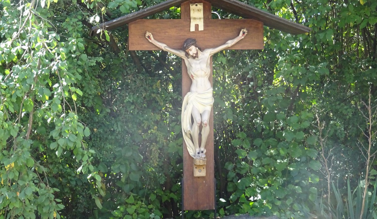 Ein Kruzifix mit einer Jesusfigur steht vor dichtem grünen Laub. Das Kreuz ist aus Holz und hat ein kleines Dach., © Foto: Cornelia Steinbach Ein Kruzifix mit einer Jesusfigur steht vor dichtem grünen Laub. Das Kreuz ist aus Holz und hat ein kleines Dach., © Foto: Cornelia Steinbach