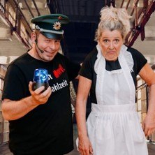 Zwei Personen in einem humorvollen Setting: Eine trägt eine Polizeimütze und hält ein blaues Licht, die andere eine Schürze. Hintergrund erinnert an Gefängnis., © links im Bild