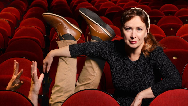 Frau sitzt in einem Theatersaal, während Beine und Hände einer anderen Person aus den roten Sitzen ragen. Humorvolle Szene., © Mauerwerk GmbH