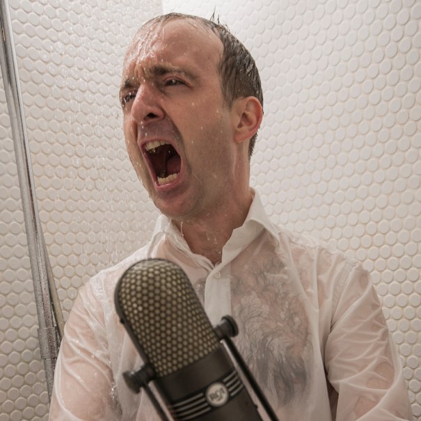 Ein Mann in einem wei&szlig;en Hemd singt leidenschaftlich unter der Dusche. Er h&auml;lt ein Mikrofon vor sich, w&auml;hrend Wasser auf ihn herabstr&ouml;mt., &copy; Scala Kultur Live gGmbH