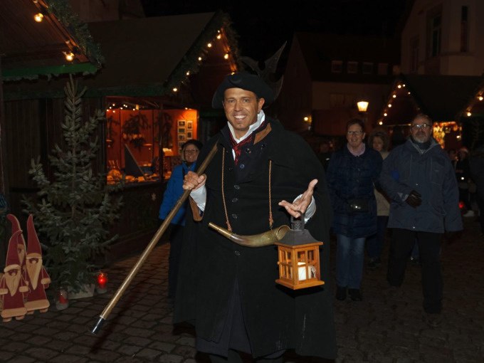 Ein Nachtw&auml;chter in traditioneller Kleidung mit Laterne und Horn f&uuml;hrt eine Gruppe von Menschen &uuml;ber einen festlich beleuchteten Weihnachtsmarkt., &copy; Stadt N&uuml;rtingen