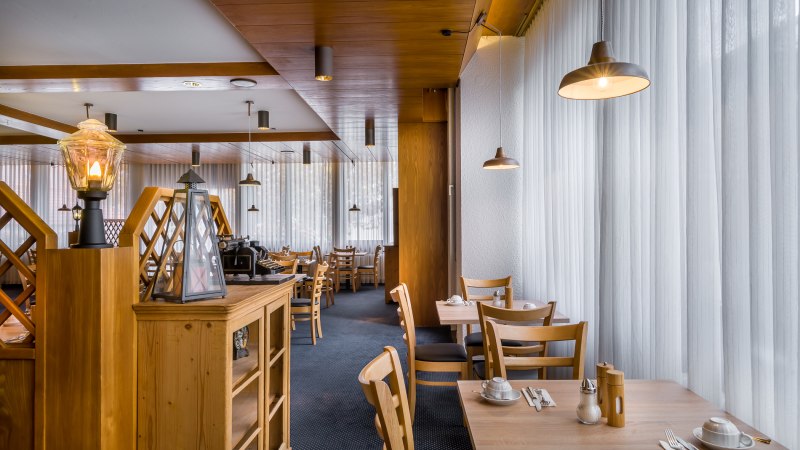 Ein Frühstücksbereich mit Holzmöbeln, gedeckten Tischen und warmem Licht. Große Fenster mit Vorhängen lassen Tageslicht herein., © Novum Management GmbH Ein Frühstücksbereich mit Holzmöbeln, gedeckten Tischen und warmem Licht. Große Fenster mit Vorhängen lassen Tageslicht herein., © Novum Management GmbH