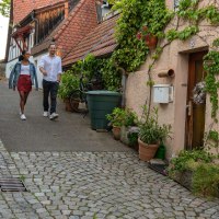 Ein Paar spaziert durch eine malerische Altstadtstra&szlig;e in Besigheim. Fachwerkh&auml;user und Pflanzen schm&uuml;cken den Weg. Weitere Personen im Hintergrund., &copy; SMG, Martina Denker