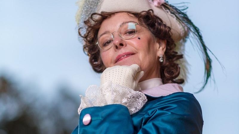 Eine Frau in historischer Kleidung mit einem verzierten Hut und Brille posiert selbstbewusst vor einem blauen Himmel., © Quelle: Irma Beck Eine Frau in historischer Kleidung mit einem verzierten Hut und Brille posiert selbstbewusst vor einem blauen Himmel., © Quelle: Irma Beck