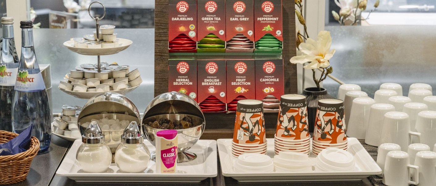 Ein Frühstücksbuffet mit verschiedenen Teesorten, Wasserflaschen, Zucker, Sahne und Kaffeebechern auf einem Tisch arrangiert., © Plaza INN Stuttgart-Filderstadt Ein Frühstücksbuffet mit verschiedenen Teesorten, Wasserflaschen, Zucker, Sahne und Kaffeebechern auf einem Tisch arrangiert., © Plaza INN Stuttgart-Filderstadt