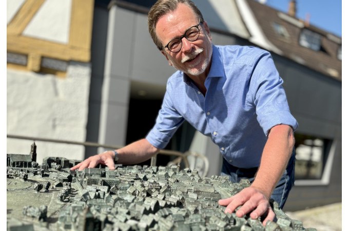 Ein Mann in einem blauen Hemd beugt sich lächelnd über ein detailliertes Stadtmodell. Im Hintergrund sind Gebäude und blauer Himmel zu sehen., © WTM GmbH Waiblingen Ein Mann in einem blauen Hemd beugt sich lächelnd über ein detailliertes Stadtmodell. Im Hintergrund sind Gebäude und blauer Himmel zu sehen., © WTM GmbH Waiblingen