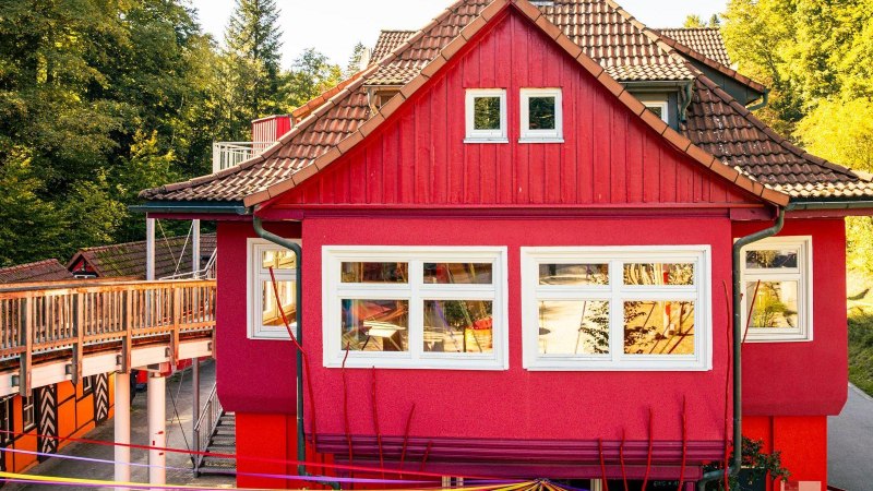 Ein rotes Gebäude mit weißen Fenstern, umgeben von grünen Bäumen. Ein Holzsteg führt am Gebäude vorbei., © Stuttgart-Marketing GmbH, Sarah Schmid Ein rotes Gebäude mit weißen Fenstern, umgeben von grünen Bäumen. Ein Holzsteg führt am Gebäude vorbei., © Stuttgart-Marketing GmbH, Sarah Schmid