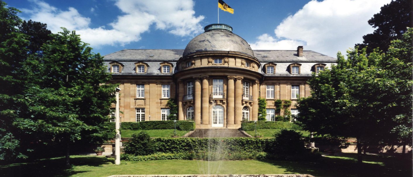 Die Villa Reitzenstein mit imposanter Fassade, einem Brunnen im Vordergrund und einer Flagge auf dem Dach, umgeben von Bäumen., © Staatsministerium Baden-Württemberg