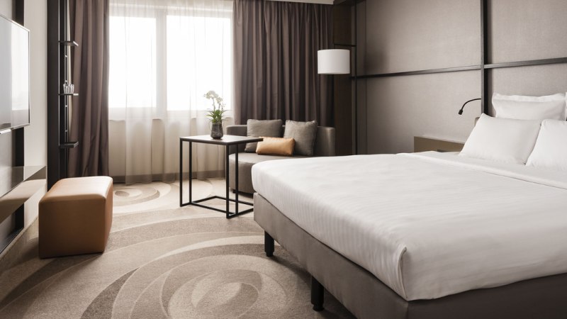 Modernes Hotelzimmer mit gro&szlig;em Bett, Sofa, Tisch und Fernseher. Helle Vorh&auml;nge und dezente Farben schaffen eine elegante Atmosph&auml;re., &copy; Stuttgart Marriott Hotel Sindelfingen