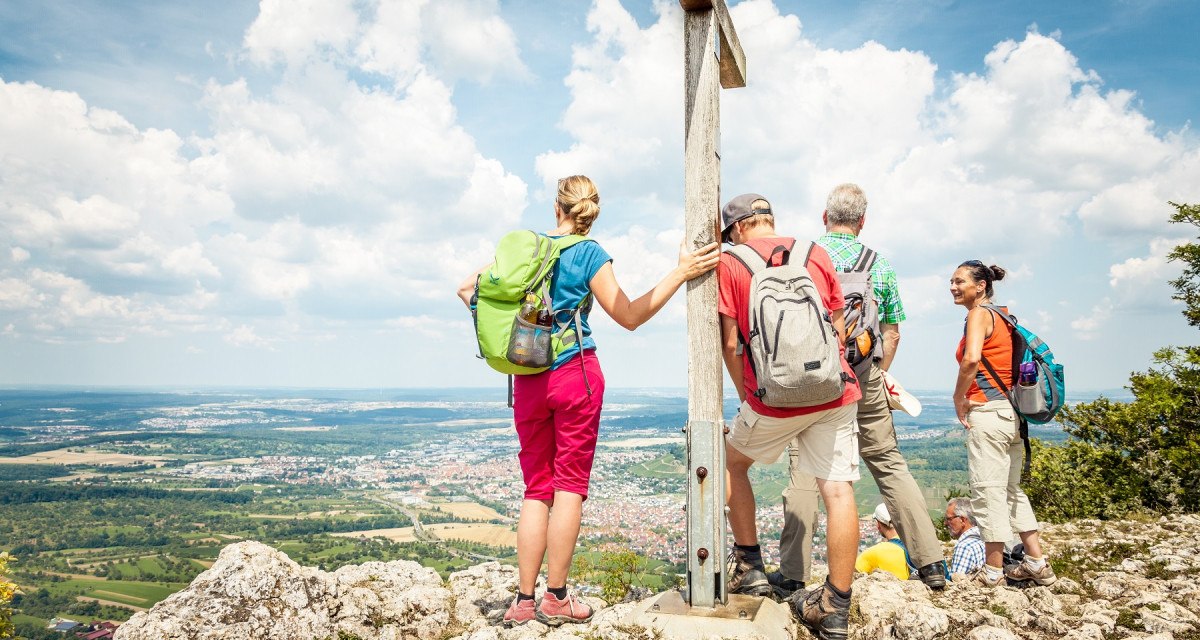 Wanderer stehen auf einem Berggipfel neben einem Gipfelkreuz und genießen die Aussicht auf die Landschaft unter einem blauen Himmel mit Wolken., © hochgehberge